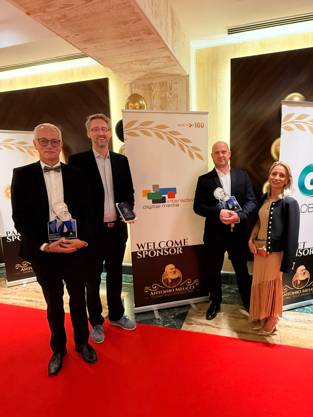 Four people holding awards on a red carpet beside a sponsor banner